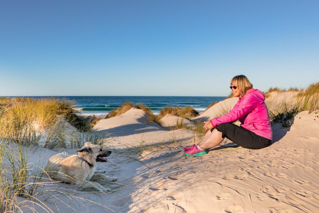 Leben im Wohnmobil Frau sitzt am Strand und schaut Ihren Hund an. Sie sitzen in den Dünen und hinter ihnen ist die Strandkulisse von der Halbinsel Peniche in Portugal